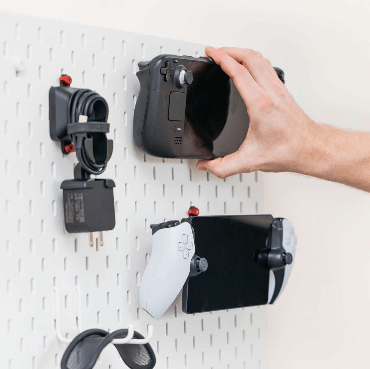 A hand attaches a Steam Deck to an Ikea Skadis Pegboard using the Mechanism Deckmate Grip and Mechanism Skadis Mount